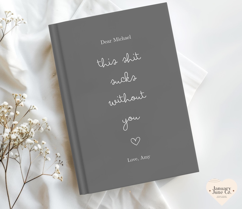 Peut inclure: Un journal gris avec le texte "Dear Michael, this shit sucks without you, Love, Amy" en &eacute;criture blanche. Un petit c&oelig;ur blanc est en dessous du texte. Le journal est sur une surface blanche avec des fleurs blanches.