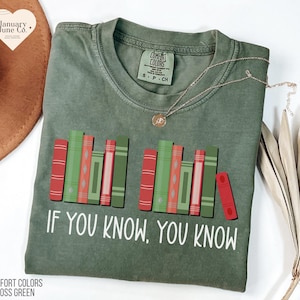 May include: Moss green Comfort Colors t-shirt with a graphic of colorful books and the text "IF YOU KNOW, YOU KNOW" in white. The shirt has a crew neck and short sleeves. A gold necklace is draped on the shirt.