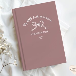 May include: A dusty rose-colored book with the title "my little book of prayers" in white script, featuring a bow illustration and the name "ELISABETH ROSE." A small heart is below the name. The book is on a white fabric surface.
