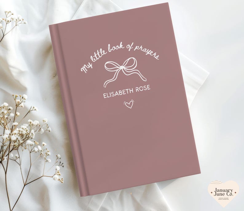 May include: A dusty rose-colored book with the title "my little book of prayers" in white script, featuring a bow illustration and the name "ELISABETH ROSE." A small heart is below the name. The book is on a white fabric surface.