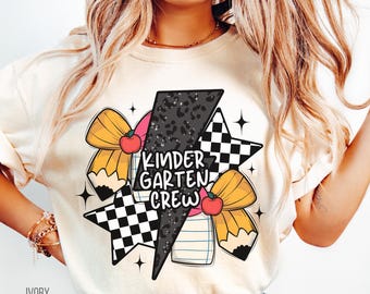 May include: A white t-shirt with a graphic design featuring a black and white checkered star, a black and white leopard print lightning bolt, a yellow bow with a red apple, and a pencil with a yellow bow. The text "KINDERGARTEN CREW" is printed in black on the lightning bolt. The t-shirt is labeled "IVORY".