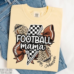 May include: Butter yellow t-shirt with a graphic that says "FOOTBALL mama" in white. The graphic includes footballs, a bow, and a lightning bolt. The shirt also has the text "#45 Johnson".