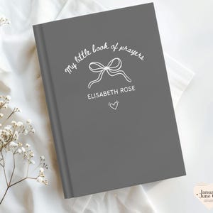 May include: A gray book with the text "my little book of prayers" and the name "ELISABETH ROSE" in white script. A bow and heart are also featured on the cover. The book is on a white surface with floral accents.
