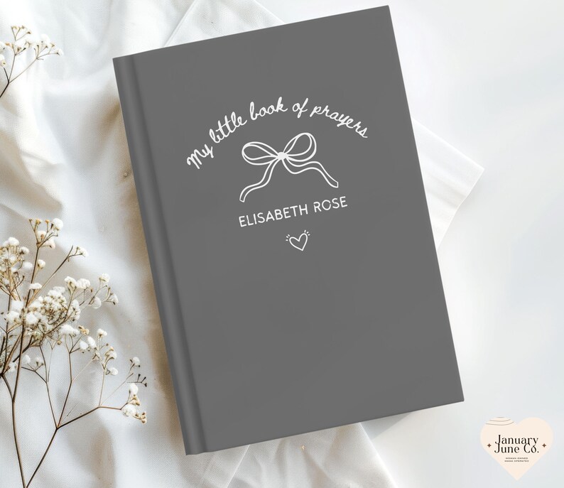 May include: A gray book with the text "my little book of prayers" and the name "ELISABETH ROSE" in white script. A bow and heart are also featured on the cover. The book is on a white surface with floral accents.