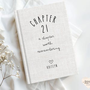 May include: A hardcover book with a light beige linen cover. The cover features the text "CHAPTER 21" in a whimsical font, followed by "a chapter worth remembering" and the name "KAITLYN" below a heart symbol. The book is a keepsake.