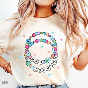 Puede incluir: Camiseta de color marfil con un dise&ntilde;o de pulsera de cuentas coloridas y el texto "MRS JOHNSON" en letras blancas. El dise&ntilde;o incluye estrellas y corazones en colores pastel. Una prenda informal.