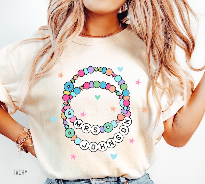 Puede incluir: Camiseta de color marfil con un dise&ntilde;o de pulsera de cuentas coloridas y el texto "MRS JOHNSON" en letras blancas. El dise&ntilde;o incluye estrellas y corazones en colores pastel. Una prenda informal.