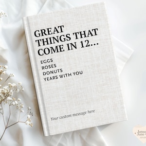 Puede incluir: Un libro de tapa dura con una cubierta de textura de lino beige claro con texto en negro que dice: GREAT THINGS THAT COME IN 12... EGGS, ROSES, DONUTS, YEARS WITH YOU, y Your custom message here. El libro está sobre una tela blanca.