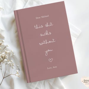 Peut inclure: Un journal mauve avec le texte "Dear Michael, this shit sucks without you" en &eacute;criture blanche. Un petit c&oelig;ur et les mots "Love, Amy" sont en dessous. Le journal est sur une surface blanche.