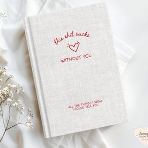 May include: A hardback journal with a light beige linen-textured cover. The cover features red text that reads "this shit sucks without you" with a heart symbol. Below, it says "ALL THE THINGS I WISH I COULD TELL YOU".
