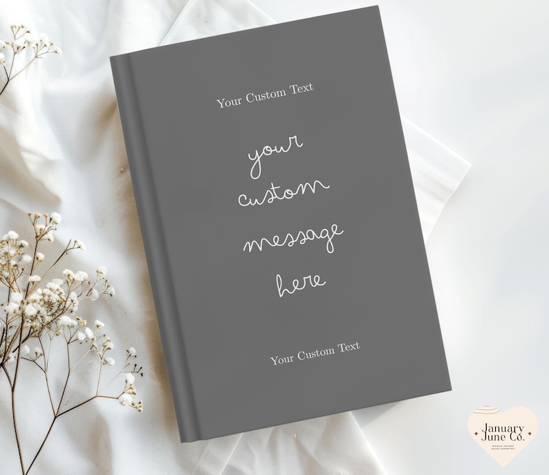 Puede incluir: Un diario de tapa dura gris con el texto "Your Custom Text" en la parte superior e inferior. Las palabras "your custom message here" est&aacute;n escritas en cursiva. El diario est&aacute; sobre una tela blanca con flores blancas.