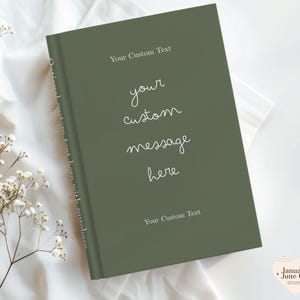 May include: A hardcover journal in a muted green color, with the text "Your Custom Text" at the top and bottom. The words "your custom message here" are written in a cursive font. The journal is on a white fabric surface.