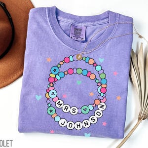Puede incluir: Camiseta violeta Comfort Colors con un dise&ntilde;o de cuentas de colores. El dise&ntilde;o incluye el texto "MRS JOHNSON" en letras blancas. Un collar dorado est&aacute; drapeado sobre la camisa, y la palabra "VIOLET" est&aacute; impresa en la parte inferior izquierda.