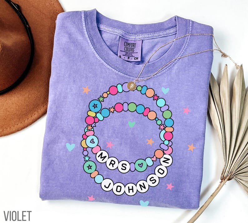Puede incluir: Camiseta violeta Comfort Colors con un dise&ntilde;o de cuentas de colores. El dise&ntilde;o incluye el texto "MRS JOHNSON" en letras blancas. Un collar dorado est&aacute; drapeado sobre la camisa, y la palabra "VIOLET" est&aacute; impresa en la parte inferior izquierda.