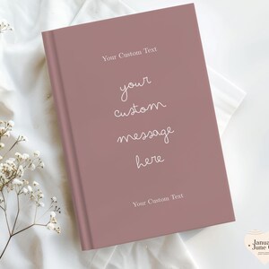 Puede incluir: Un diario rosa polvoriento con el texto "Your Custom Text" en la parte superior e inferior, y "your custom message here" en el centro. El diario est&aacute; cerrado y descansa sobre una superficie de tela blanca, con delicadas flores blancas en la esquina.