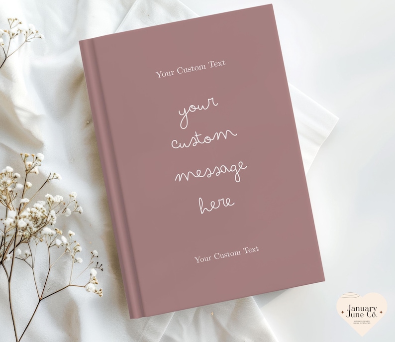 Puede incluir: Un diario rosa polvoriento con el texto "Your Custom Text" en la parte superior e inferior, y "your custom message here" en el centro. El diario est&aacute; cerrado y descansa sobre una superficie de tela blanca, con delicadas flores blancas en la esquina.