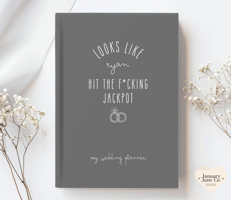 K&ouml;nnte beinhalten: Ein graues Hochzeitsplaner-Notizbuch mit wei&szlig;em Text, der "LOOKS LIKE ryan HIT THE F*CKING JACKPOT" und "my wedding planner" lautet. Das Design beinhaltet zwei Eheringe. Ein Hochzeitsplanungsartikel.
