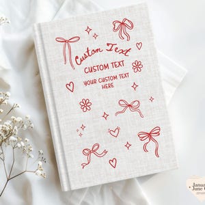 May include: A hardcover journal with a light beige linen cover. Red illustrations of bows, hearts, flowers, and stars decorate the cover. The words "Custom Text" are written in red script, with space for personalized text.