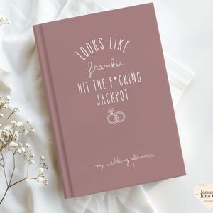 K&ouml;nnte beinhalten: Ein staubrosa Hochzeitsplaner mit wei&szlig;em Text. Auf dem Cover steht "Looks like Frankie hit the f*cking jackpot" mit einer Ringgrafik und "my wedding planner" darunter.