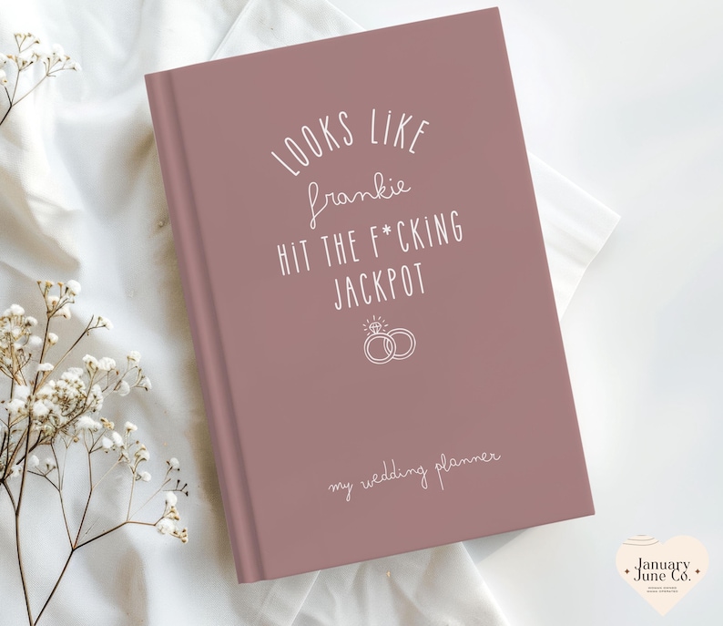 K&ouml;nnte beinhalten: Ein staubrosa Hochzeitsplaner mit wei&szlig;em Text. Auf dem Cover steht "Looks like Frankie hit the f*cking jackpot" mit einer Ringgrafik und "my wedding planner" darunter.