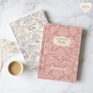 May include: Two hardcover journals with decorative patterns and the text "CHAPTER thirty." One journal is cream-colored with blue illustrations, the other is pink with cream illustrations. A cup of coffee and flowers are in the background.