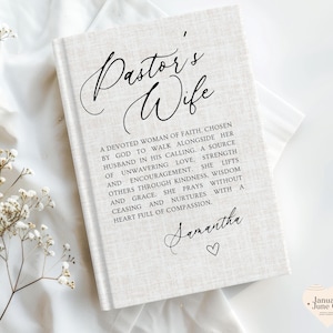 Puede incluir: Un libro de tapa dura con una cubierta de textura de lino beige claro. El frente presenta el texto negro Pastor's Wife en cursiva, seguido de un párrafo que describe a una mujer de fe, el nombre Samantha en cursiva y un pequeño corazón.