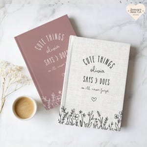 May include: Two hardcover journals, one dusty rose and one off-white, with the text "CUTE THINGS olivia SAYS & DOES so ill never forget." Floral line art decorates the bottom of the covers. A cup of coffee sits nearby.