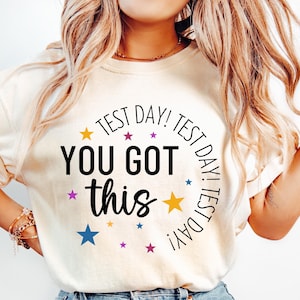 May include: White t-shirt with a black circle graphic that says "Test Day! Test Day! Test Day! You Got This". The graphic is surrounded by colorful stars.