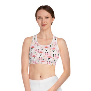May include: White sports bra with a repeating pattern of ghost faces wearing pink cowboy hats and boots, with cacti and stars on a light pink background. The bra has a scoop neck and racerback design.