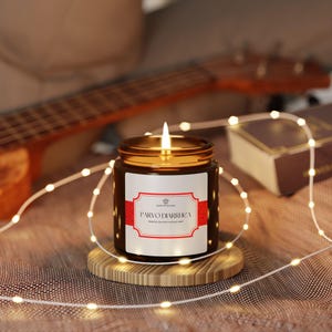 May include: A burning candle in a dark glass jar with a gold lid, on a wooden coaster. The label says "PARVO DIARRHEA" in red and white. Fairy lights and a ukulele are in the background.