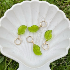 May include: Four green glass leaf charms with gold wire loops. The charms are on a white seashell-shaped dish.
