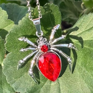 May include: A silver-toned spider pendant necklace with a large, faceted red gemstone body and smaller red gem accents. The spider has detailed legs and is suspended from a delicate chain. The necklace is displayed against a backdrop of green leaves.