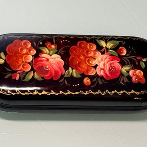 May include: A black, oval-shaped box with a glossy finish, adorned with a floral design. The flowers are painted in shades of red and pink, with green leaves. A gold-colored decorative border lines the edge of the box.