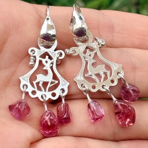 May include: A pair of silver dangle earrings featuring a cutout deer silhouette within a decorative frame. Each earring is accented with three small, irregular, translucent pink gemstone drops hanging from the bottom edge of the metal design.