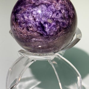 May include: A polished sphere of charoite, displaying swirling patterns of deep purple and lavender hues. The gemstone sits on a clear, petal-shaped stand, creating a floating effect. This decorative item is ideal for home decor.