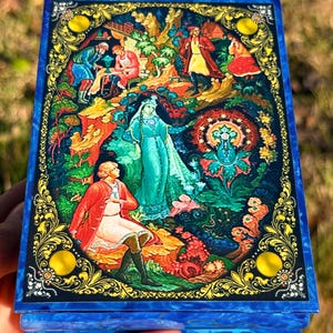 May include: A blue lacquered box with a detailed, colorful illustration. The artwork depicts a scene with figures in traditional clothing, surrounded by foliage and ornate gold filigree. The box has a glossy finish.