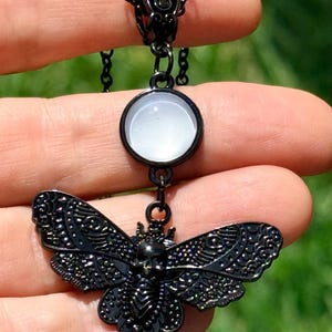 May include: A black metal necklace featuring a circular pendant with a smooth white cabochon stone, connected to a detailed moth-shaped charm. The moth charm has intricate wing patterns and a small skull design at the center, suspended on a chain.