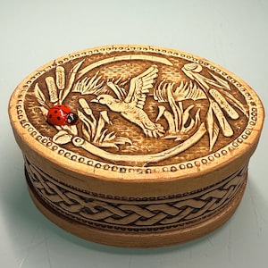 May include: Oval wooden box with a carved relief design. The lid features a flying bird, cattails, and a red ladybug. The sides of the box have a woven pattern. The box is made of light brown wood.