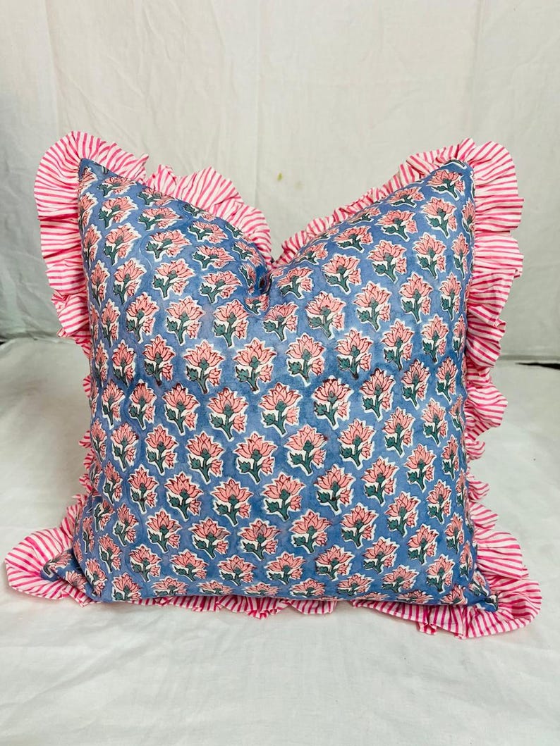 May include: A square throw pillow featuring a blue fabric base with a repeating pattern of pink flowers and green leaves. The pillow is bordered by a ruffled edge made of fabric with thin, alternating white and pink vertical stripes.