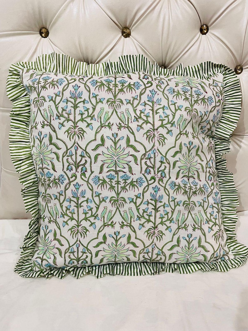 May include: A square throw pillow featuring a white base with a repeating pattern of green vines, light blue flowers, and small green birds. The pillow is finished with a ruffled border made of fabric with thin green and white vertical stripes.