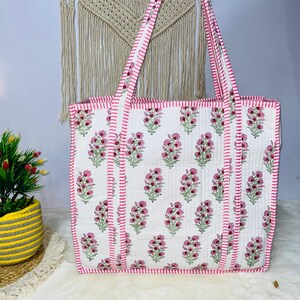 May include: A white cotton tote bag with a pink floral print and pink and white striped straps. The bag has a quilted texture and a pink stitched border.