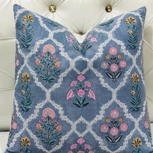 K&ouml;nnte beinhalten: Ein quadratisches Zierkissen mit einem staubblauen Stoffgrund und einem wei&szlig;en Gittermuster. Das Design zeigt sich wiederholende florale Motive in Rosa-, Gelb- und Gr&uuml;nt&ouml;nen, die innerhalb jedes Abschnitts des wei&szlig;en dekorativen Gitters angeordnet sind.