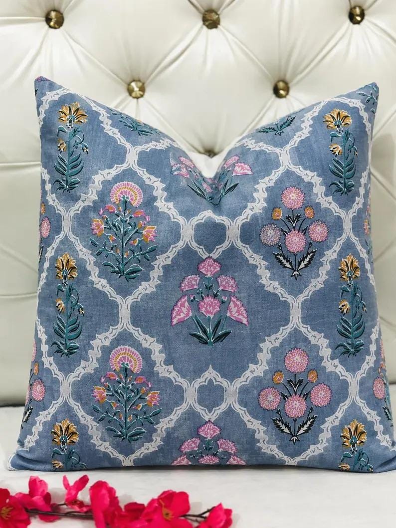 K&ouml;nnte beinhalten: Ein quadratisches Zierkissen mit einem staubblauen Stoffgrund und einem wei&szlig;en Gittermuster. Das Design zeigt sich wiederholende florale Motive in Rosa-, Gelb- und Gr&uuml;nt&ouml;nen, die innerhalb jedes Abschnitts des wei&szlig;en dekorativen Gitters angeordnet sind.