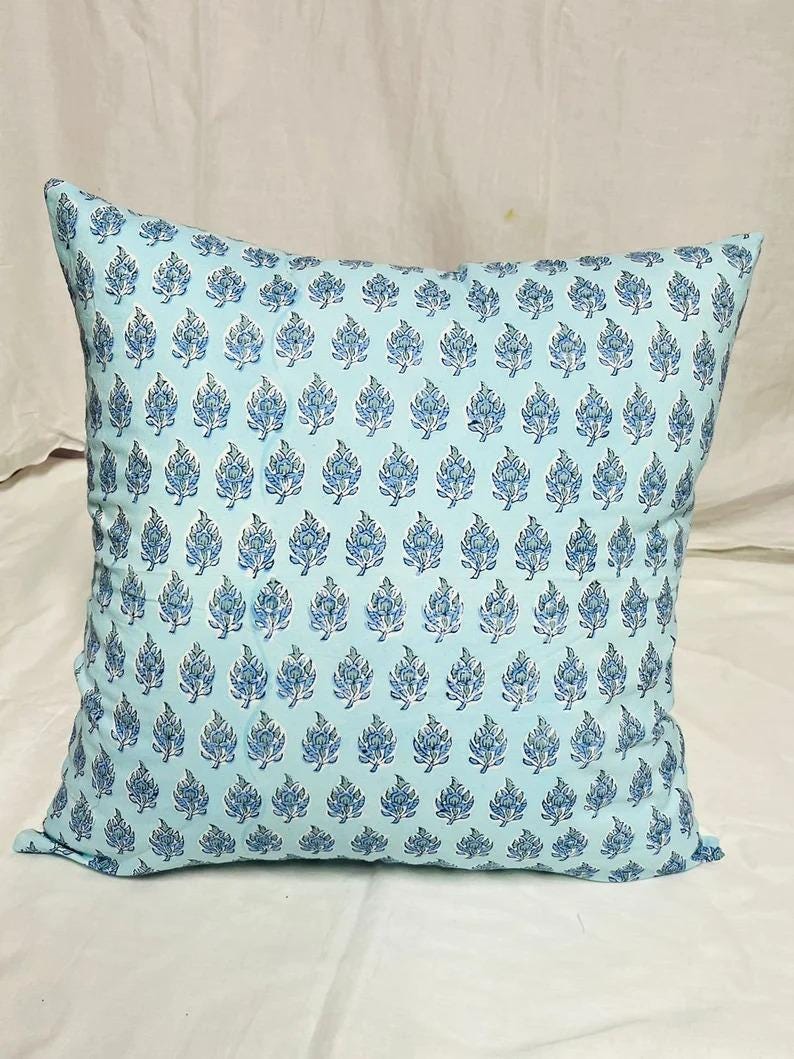 K&ouml;nnte beinhalten: Ein quadratisches Dekokissen mit hellblauem Hintergrund und einem sich wiederholenden Muster aus dunkelblauen Blumenmotiven. Das Design besteht aus kleinen, stilisierten Blatt- und Bl&uuml;tenformen, die in gleichm&auml;&szlig;igen Reihen angeordnet sind.