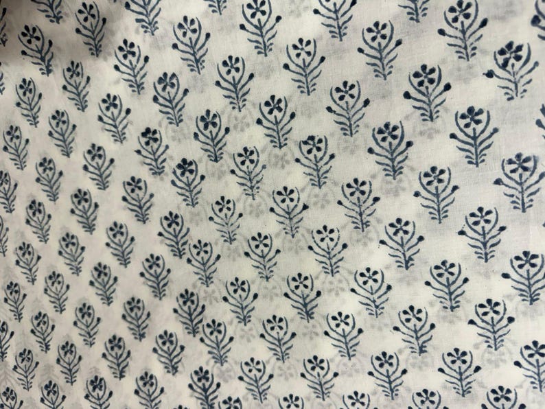 K&ouml;nnte beinhalten: Ein wei&szlig;er Stoff mit einem sich wiederholenden Muster aus kleinen, dunkelblauen Blumenmotiven. Jedes Motiv besteht aus einem zentralen Stiel mit symmetrischen Bl&auml;ttern und einer f&uuml;nfbl&auml;ttrigen Bl&uuml;te an der Spitze, die gleichm&auml;&szlig;ig angeordnet sind.