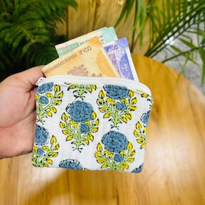 Puede incluir: Un monedero pequeño y rectangular de tela acolchada con base blanca y un patrón repetitivo de flores azules y hojas amarillas. El monedero está abierto por la parte superior, mostrando varios billetes de rupias indias en el compartimento.