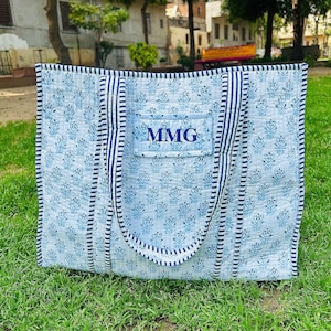 Puede incluir: Una bolsa de mano acolchada azul y blanca con un asa a rayas azul y blanca. La bolsa tiene un pequeño bolsillo en la parte delantera con las iniciales "MMG" bordadas en azul.