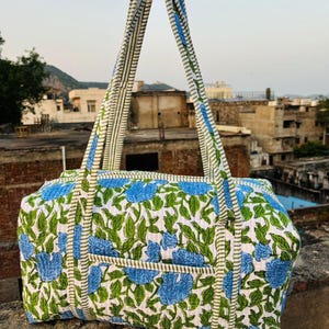 May include: A quilted duffel bag featuring a white base with a repeating pattern of blue flowers and green leaves. The bag includes two long shoulder straps with a green and white striped design, matching the trim along the bag's edges.