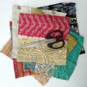 May include: A stack of colorful fabric scraps with a pair of silver scissors on top. The fabric scraps are various patterns and colors, including red, green, blue, and white.