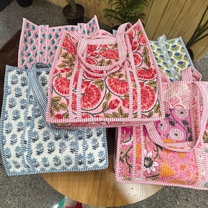 Könnte beinhalten: Eine Sammlung gesteppter Tragetaschen mit floralen Mustern. Die Taschen zeigen verschiedene Designs, darunter rosa und rote Blumen, blau-weiße Blumendrucke und rosa Hintergründe. Jede Tasche hat passende Griffe und Verzierungen.
